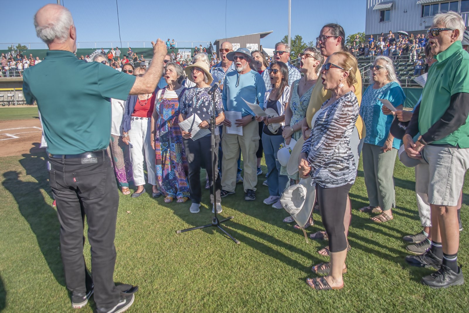 The director leading the choir on the field, singers holding sheet music in the evening sun