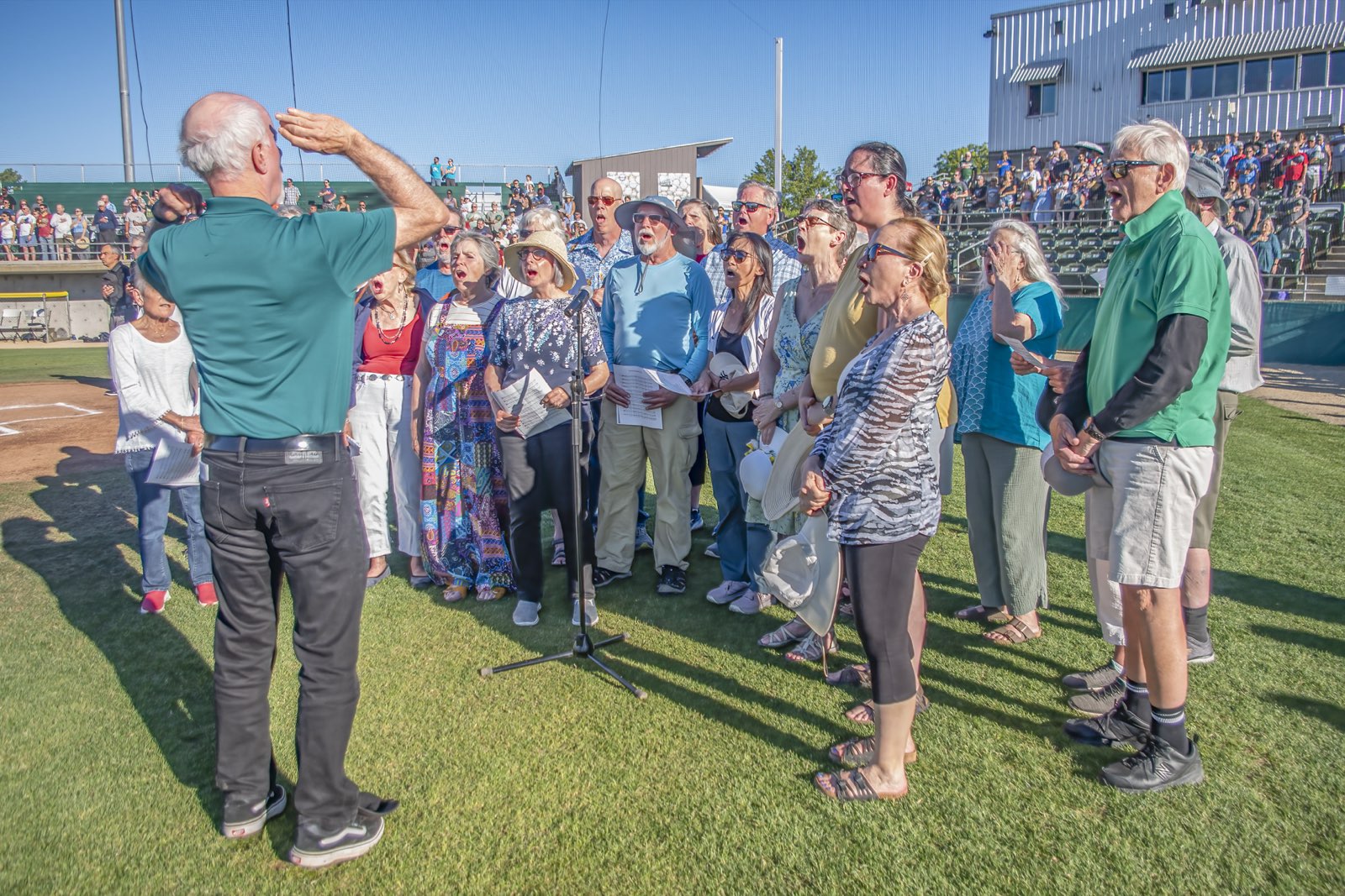 Siskiyou Singers gathered around a microphone on the infield, singing the national anthem as fans stand in the grandstand behind them